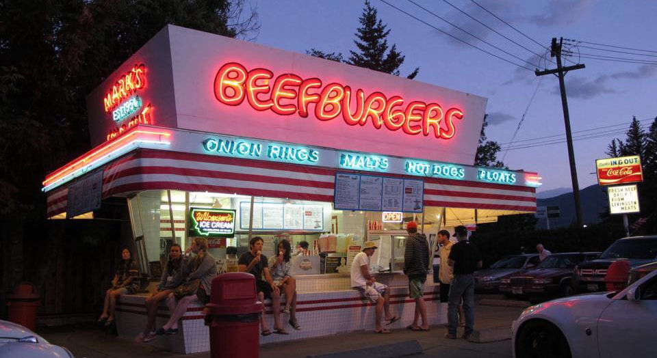 Mark's Beefburgers at dusk with full neon lights glowing and customers sitting on the bench outside the Livingston Montana drive-in