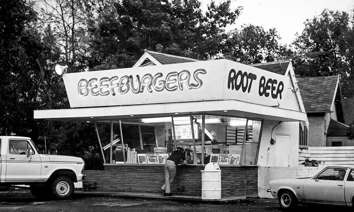 Black and white photograph of the original Mart's In and Out building with Beefburgers and Root Beer signs, vintage trucks parked in front