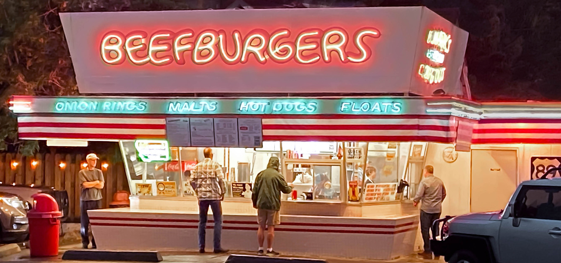 Mark's In and Out Beefburgers drive-in at night with glowing neon BEEFBURGERS sign and customers at the walk-up window in Livingston Montana